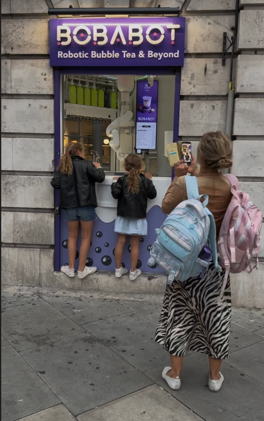 Two girls press up against the BobaBot kiosk window under the lit sign while their mum films the robot on her phone.